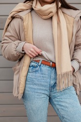 Young Girl model in fashion outerwear look with knitted vintage sweater, beige Down-padded coat jacket, scarf, blue jeans and leather belt. Women's autumn-winter stylish clothes 