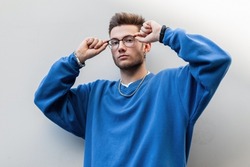 Handsome hipster guy in a trendy blue sweatshirt wears glasses and looks at the camera near the white wall in the city