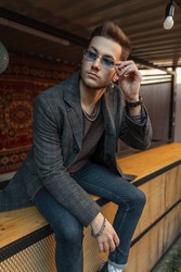 Fashionable vogue hipster man in stylish casual coat and jeans sits and wears a sunglasses in the city