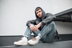 Trendy cool urban handsome man model hipster with leather jacket, hoodie, jeans and white sneakers sits on the street near a white wall