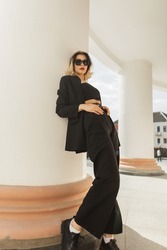 Elegant vogue beautiful young business woman model with cool trendy sunglasses in fashion strict black outfit with jacket, pants, top and shoes standing near a white vintage column in the city 