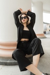 Stylish cool young woman with cool sunglasses wearing trendy black elegant clothes with a business jacket, top and pants sits and poses near a white column in town. Female vogue style and beauty 