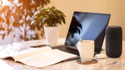 A portable speaker with built-in smart intelligence. Musical column. Marble table, work atmosphere, laptop, green flower, coffee cup. Morning sunlight.