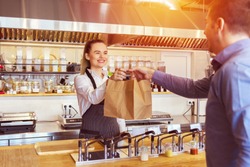 Cheerful waitress wearing apron serving customer at counter in restaurant - Small business and service concept with young woman owner offering recycled paper bag with take away food to online client
