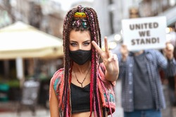 Young mixed race woman wearing face mask showing peace sign during city street protest – Hipster female protesting outdoor for human rights while people holding unity sign in background