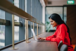 A young and attractive Asian Indian student woman works on her notebook laptop at a wooden desk during the day. She is focused and typing on her notebook; the very image of productivity.