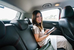 A young Japanese Asian woman sits and reads her e reader while sitting in the backseat of a car she booked via a ride hailing app. She is smiling happily as she reads and is driven to her destination.