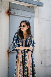 A tall, elegant, and beautiful Indian Asian woman in a summer dress and sunglasses smiles as she poses on a street in an alley during the day. She is fashionable and exudes confidence. 