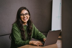 Portrait of a young, confident and attractive Indian Asian student working or studying on her laptop as she sits in a booth. She is dressed in a preppy sweater and shirt and glasses. 