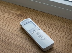 White remote control panel for motorized roller shades or automatic blinds lies on a wooden windowsill near the window.
