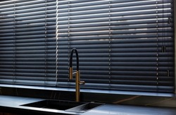 Wood blinds black color closeup on the window. Wooden slats 50mm wide. Venetian bamboo blinds in the kitchen. Black tapes. Sink with copper faucet near the window. Stone countertop.
