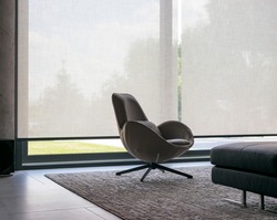 Roller blinds of large sizes on the window in the interior. Automatic solar shades, fabric with linen texture. In front of a large window is a chair on a carpet. Outside is a view of the garden.