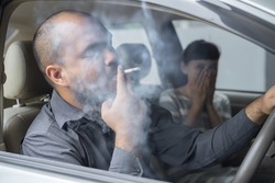 Husband smoking cigarette and wife choking of smoke. Man smoking cigarette and woman is covering her face and a lot of smoke around in car smelling pollution. Passive stop smoking cigarettes campaign.