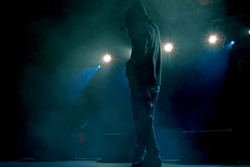 Rap artist on stage in the rays of soffits light. Concert backlight and illumination during music concert. Singer in hoodie with microphone at stage
