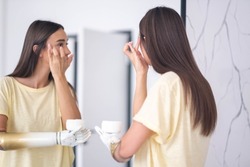woman with prosthetic arm applying cream near mirror at home, girl with disability using artificial prosthetic limb to hold cream