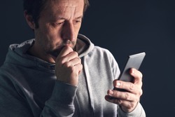 Worried casual mid-adult man looking at the screen of his smartphone with one hand on the chin