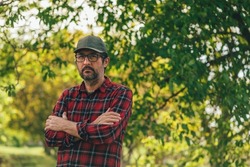 Portrait of male farmer with arms crossed wearing plaid shirt and trucker's hat posing in walnut orchard and looking at camera, selective focus