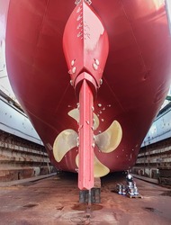 Dry-cargo ship in dry dock for repairs and maintenance at a shipyard. The stern of a large ship with a copper propeller, with a steering gear on a slipway in a dry dock.