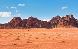 Red dusty desert with large rock massif and blue sky in background, small off road vehicle in foreground for scale. Typical landscape of Wadi Rum, Jordan