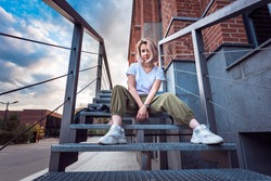 Girl in hip hop clothes sitting on a metal staircase looking at camera with a rebel look in an urban environment at sunset