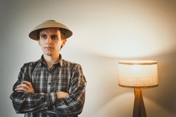 Funny photo of a guy in oriental chinese wooden hat near the handmade lamp. Man in the hat from plate is standing near the white wall. Funny and laughing person in checkered shirt on the photo.