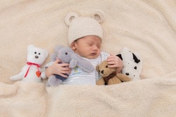 Asian newborn baby sleeping with doll, cute little toddler sleep in bed with favourite rabbit doll and teddy bear. Adorable infant wearing hat beige knitted and lying under beige blanket with toys.