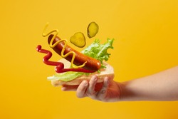 Hand holds hot dog in which sausage, salad, mustard, ketchup, pickle cucumbers are flying on a yellow background
