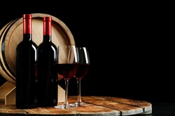 Glasses with delicious wine, bottles and barrel on table against dark background