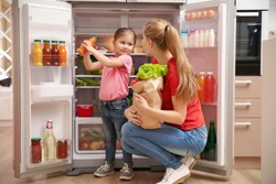 Young mother with daughter putting food into refrigerator at home after shopping