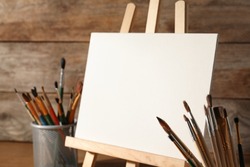 Easel with blank canvas board and brushes on table near wooden wall. Children's painting