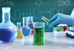 Scientist pouring liquid into beaker at table against chalkboard, closeup. Chemistry glassware