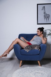 Young woman with cat and magazine on armchair at home. Cute pet