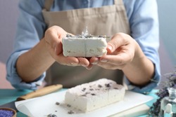 Woman holding hand made soap bar with lavender flowers at light blue wooden table, closeup
