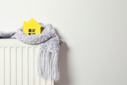 House model wrapped in scarf on radiator indoors, space for text. Winter heating efficiency