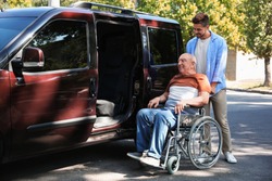 Young man helping patient in wheelchair to get into van outdoors