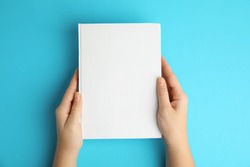 Woman holding book with blank cover on blue background, top view