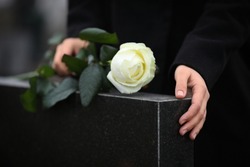Woman holding white rose near black granite tombstone outdoors, closeup. Funeral ceremony