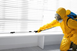 Male worker in protective suit spraying insecticide on window sill indoors. Pest control