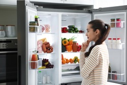 Beautiful young woman near open refrigerator in kitchen