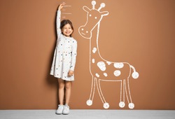 Little girl measuring height and drawing of giraffe near brown wall
