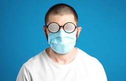 Man with foggy glasses caused by wearing disposable mask on blue background. Protective measure during coronavirus pandemic