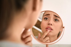 Emotional woman with herpes touching lips in front of mirror against light background