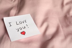 Sticky note with phrase I love you! on bed, closeup. Space for text