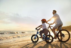 Happy father with son riding bicycles on sandy beach near sea at sunset