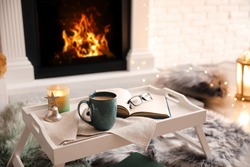 Cup of coffee, burning candle and book on tray near fireplace indoors. Cozy atmosphere