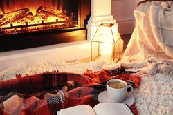 Cup of hot drink and book on blanket near fireplace indoors. Magic atmosphere