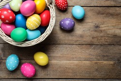 Colorful Easter eggs in wicker basket on wooden table, flat lay. Space for text