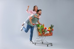 Young couple with shopping cart full of groceries on grey background