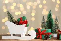 Small white sleigh, decorative Christmas trees and gift boxes on wooden table against blurred lights