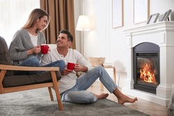 Happy couple with cups of hot drink resting near fireplace at home